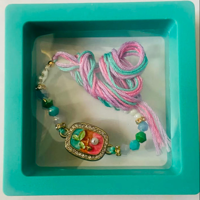 A handmade beaded bracelet with a central floral charm, resting inside a turquoise square display box. The bracelet features pink and mint green braided cords, green and white beads, and a gold-toned heart-shaped charm with a pink and orange floral design.